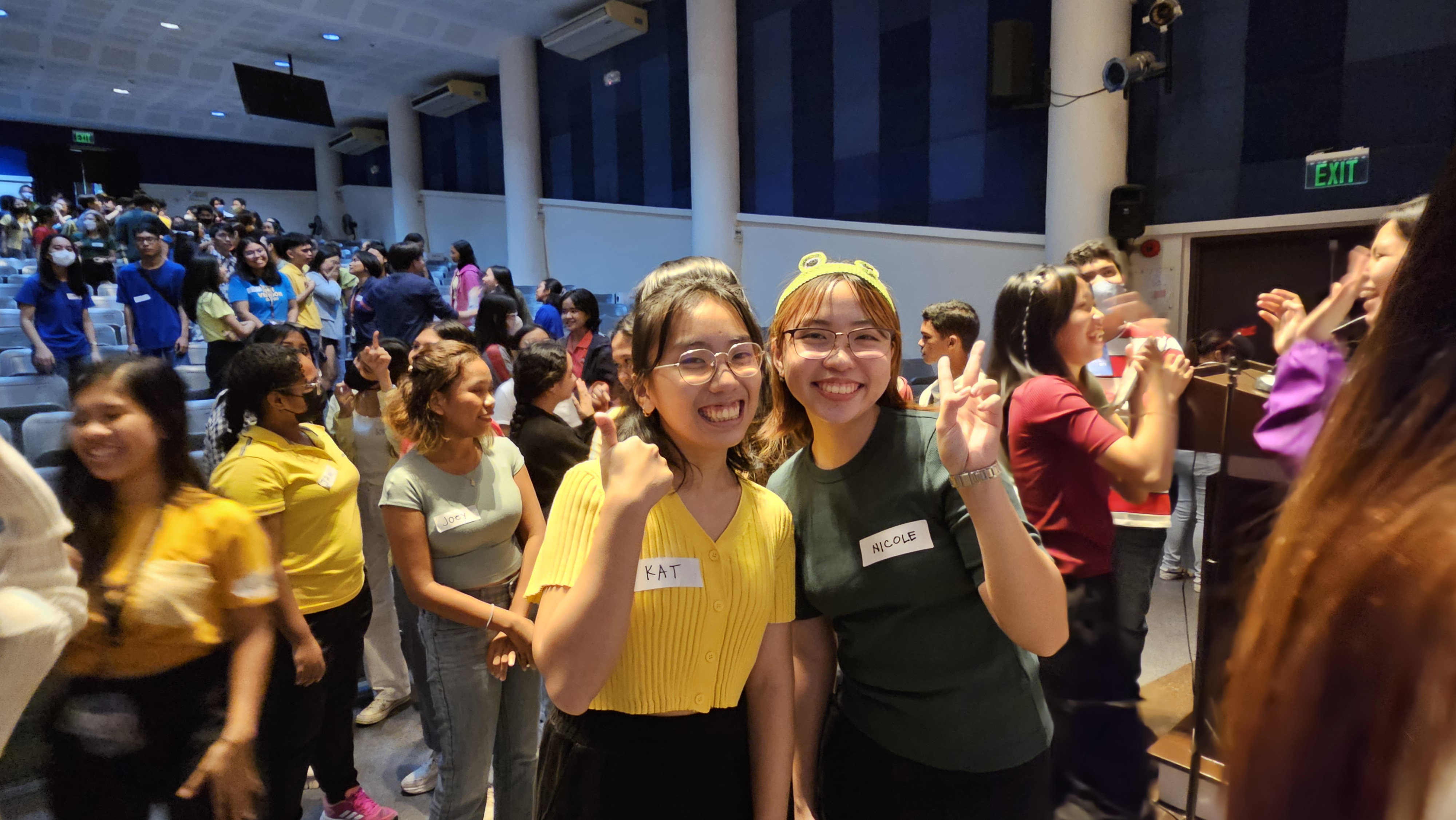 In the foreground, two students smile at the camera. On the left is one in yellow with her thumb up, on the right is one in green showing a peace sign.