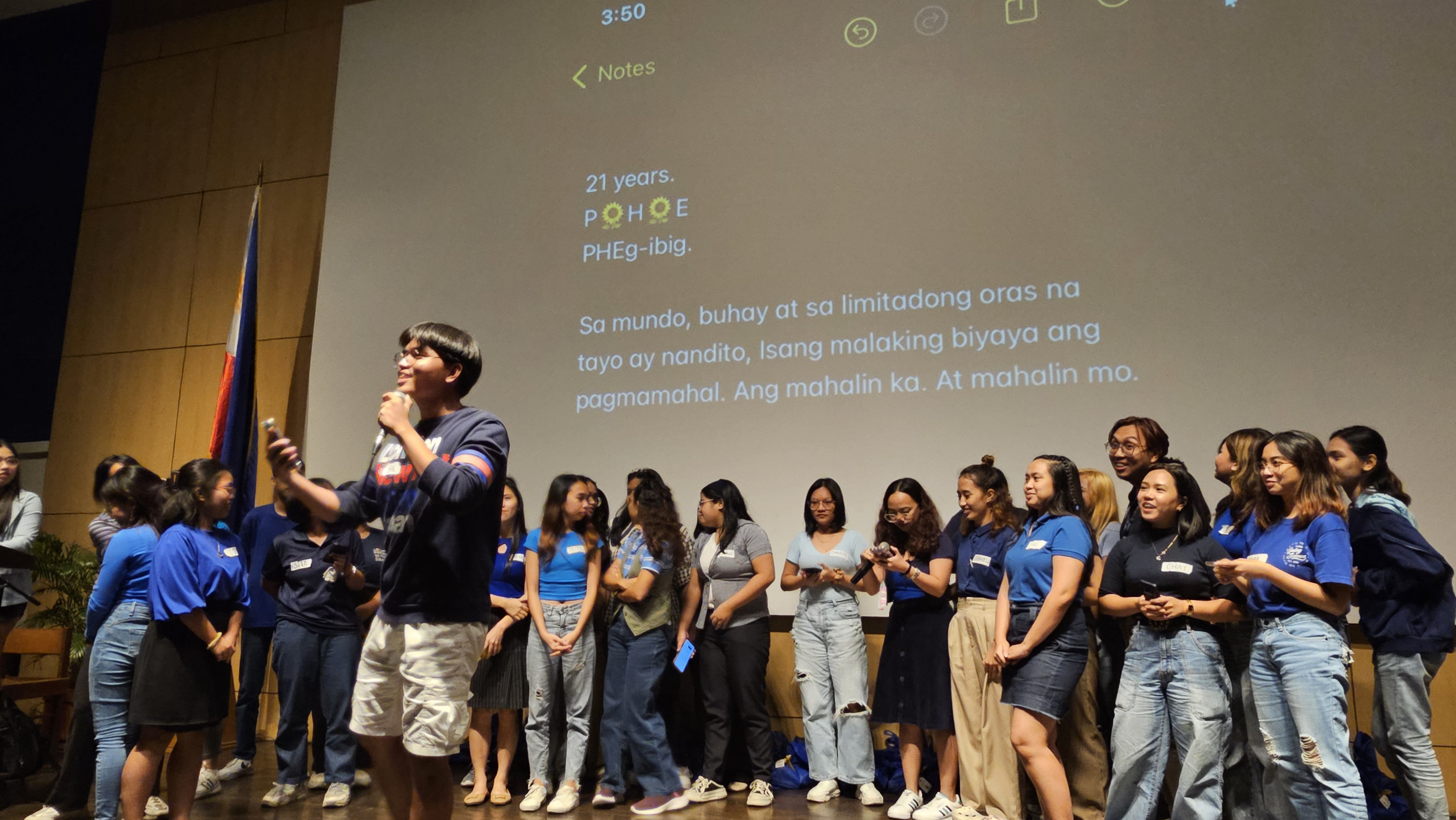 A row of students in blue stand behind one student with a microphone in his hands.