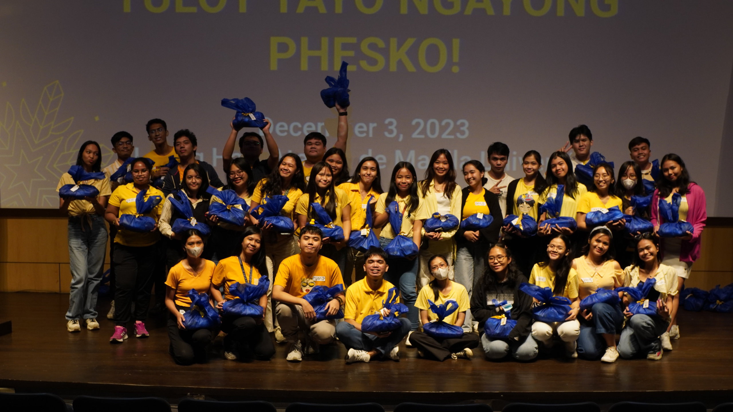 Two rows of students in yellow stand on the stage holding blue fabric bags of groceries.