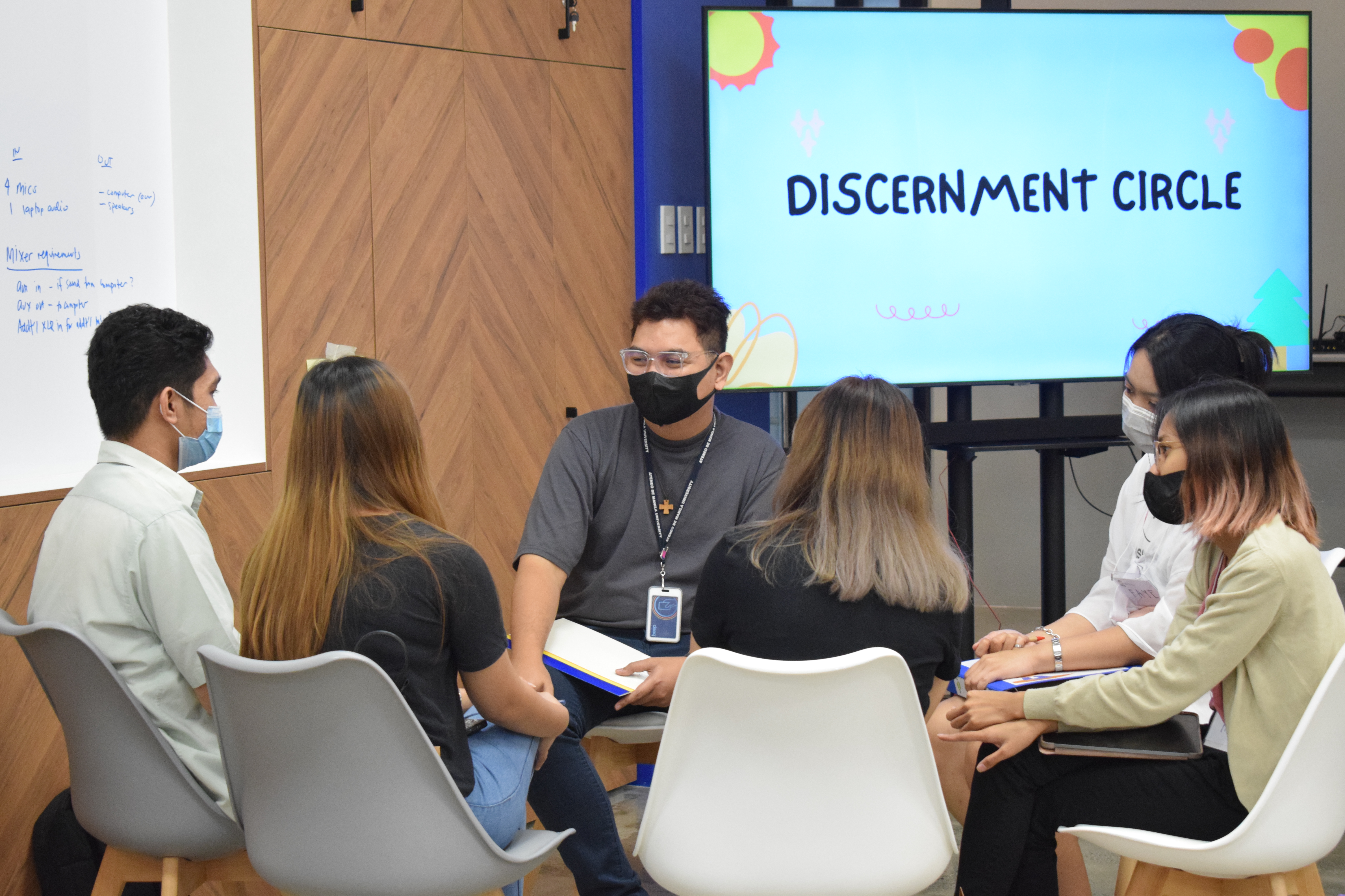 In the foreground, six people sit in a circle listening to the one speaking in the left most. In the background is a TV that says "Discernment Circles".