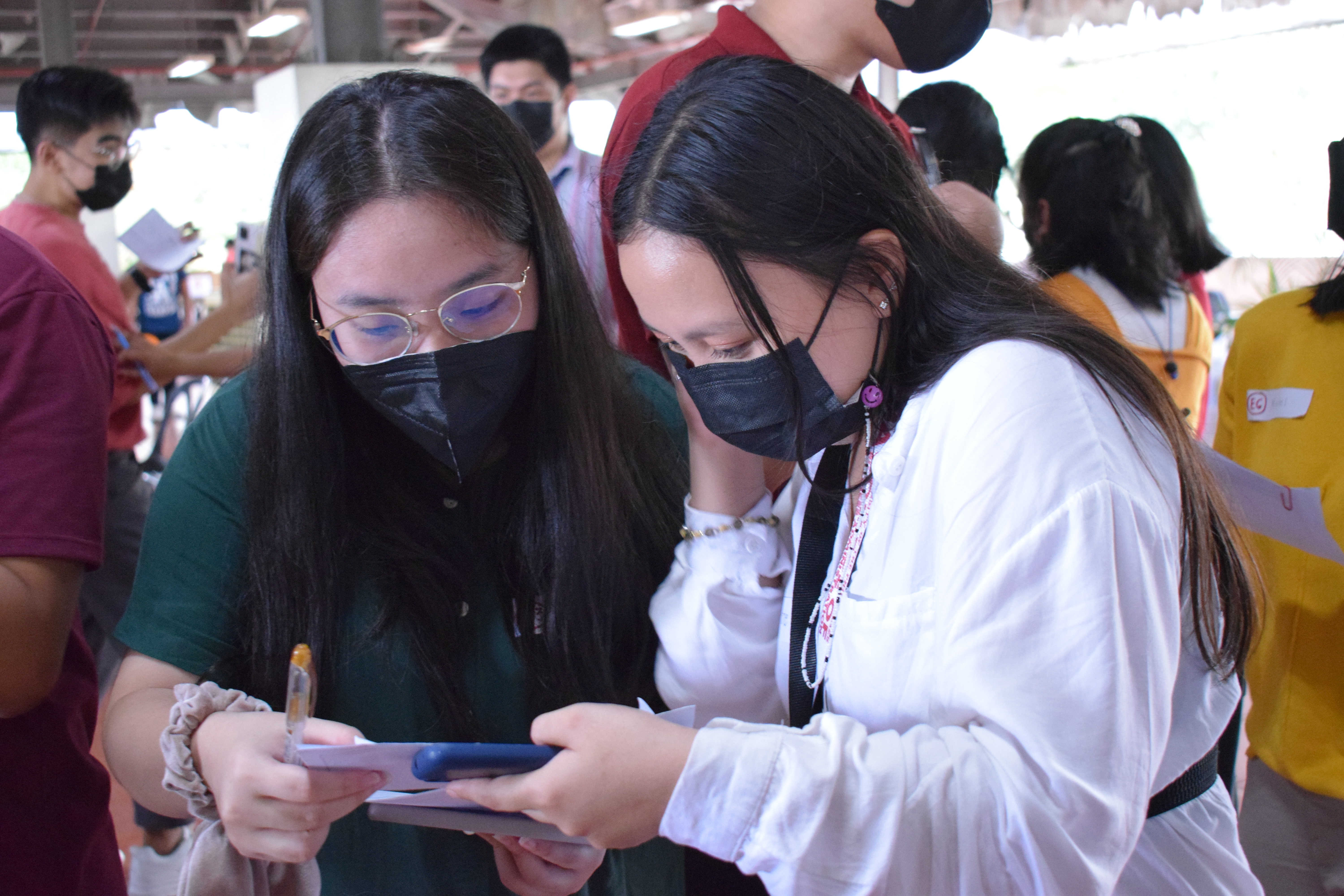 A girl in white and a girl in green look at each other's human bingo cards to see if they wrote any similar experiences. Both are wearing masks.