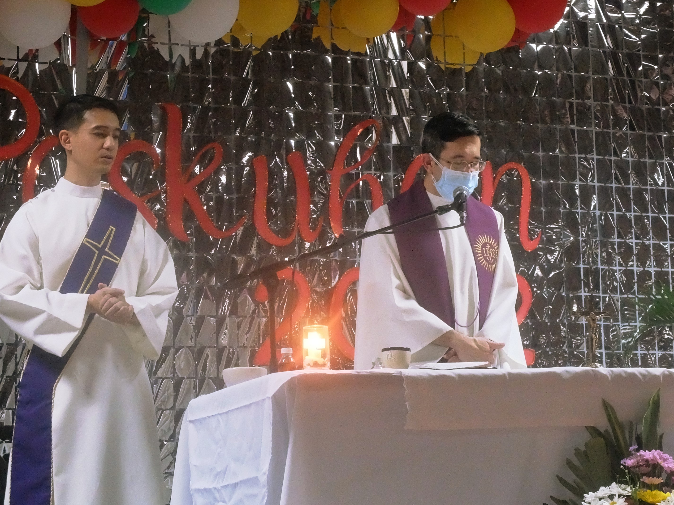 A Catholic deacon with a purple sash stands in attention to a Catholic priest in a purple stole. The priest is looking down at the altar.
