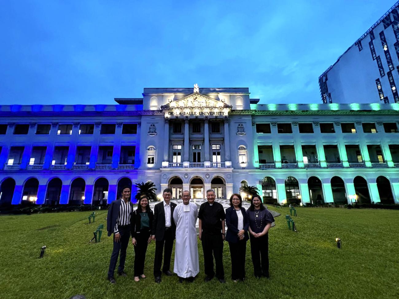 Standing before the iconic De La Salle building lit up in the colors of both schools. Left to right - Dr Norby R Salonga; Ms Fritzie Ian P De Vera; Dr Robert Roleda; Brother Bernard S Oca, FSC; Fr Roberto Yap, SJ; Dr Maria Luz Viches; Ms Ana Marina Tan