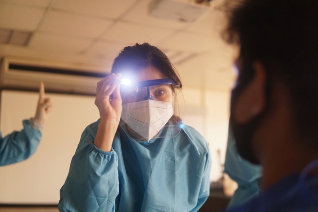 A doctor in scrubs, face mask, and face shield, shining a light onto a patient